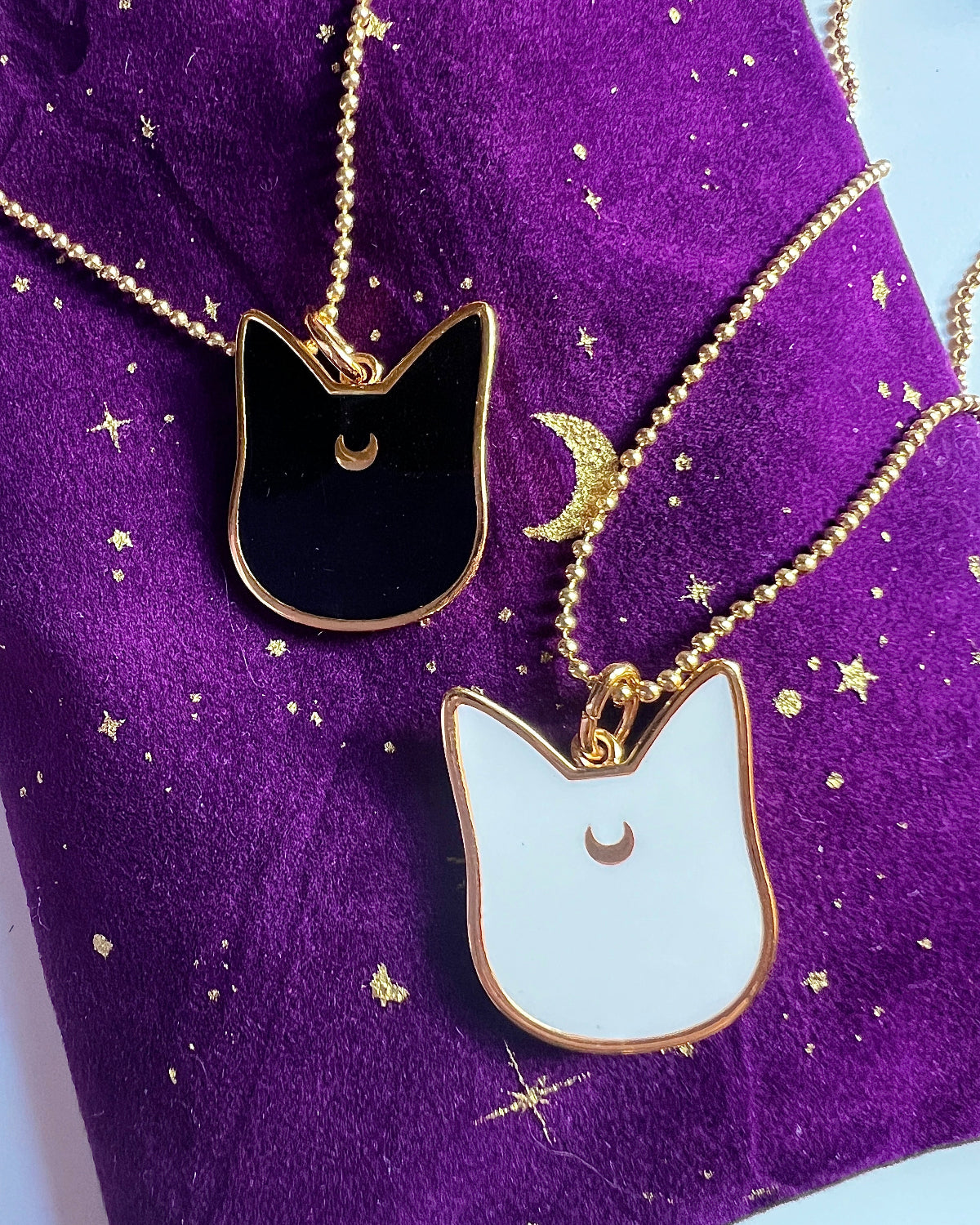 Two cat-shaped pendants on gold chains against a purple fabric background with star patterns.