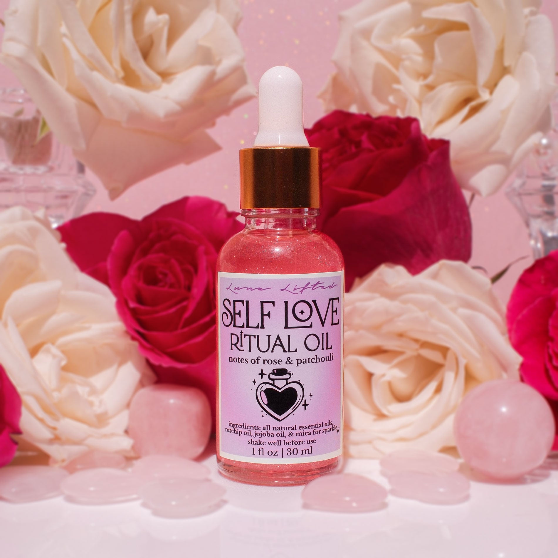 A 1 fl oz bottle of Love Lifter Self Love Ritual Oil, featuring pink rose and patchouli oil with a white dropper cap and a metallic gold collar. The bottle is centered on a white surface surrounded by pink rose petals, pink crystal balls, and fresh pink and cream roses in the background.